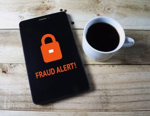 Picture of a cell phone with the words fraud alert on the screen next to a cup of coffee