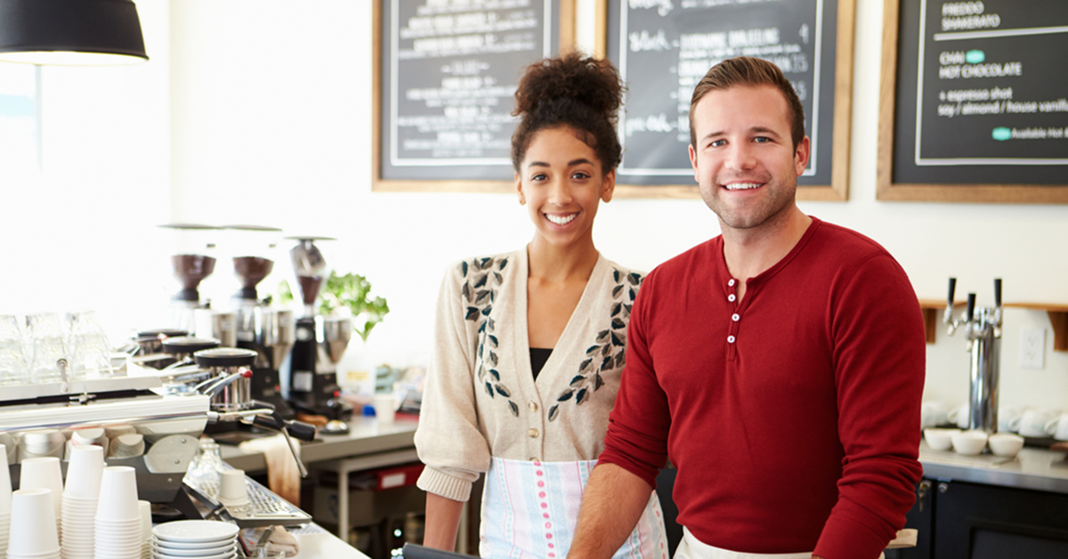 Smiling small business owners standing behind the counter of a modern coffee shop, ready to serve customers.
