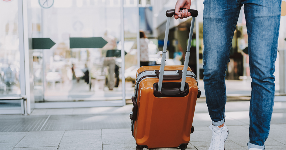 Traveler pulling an orange suitcase through airport doors, dressed casually in jeans and sneakers.