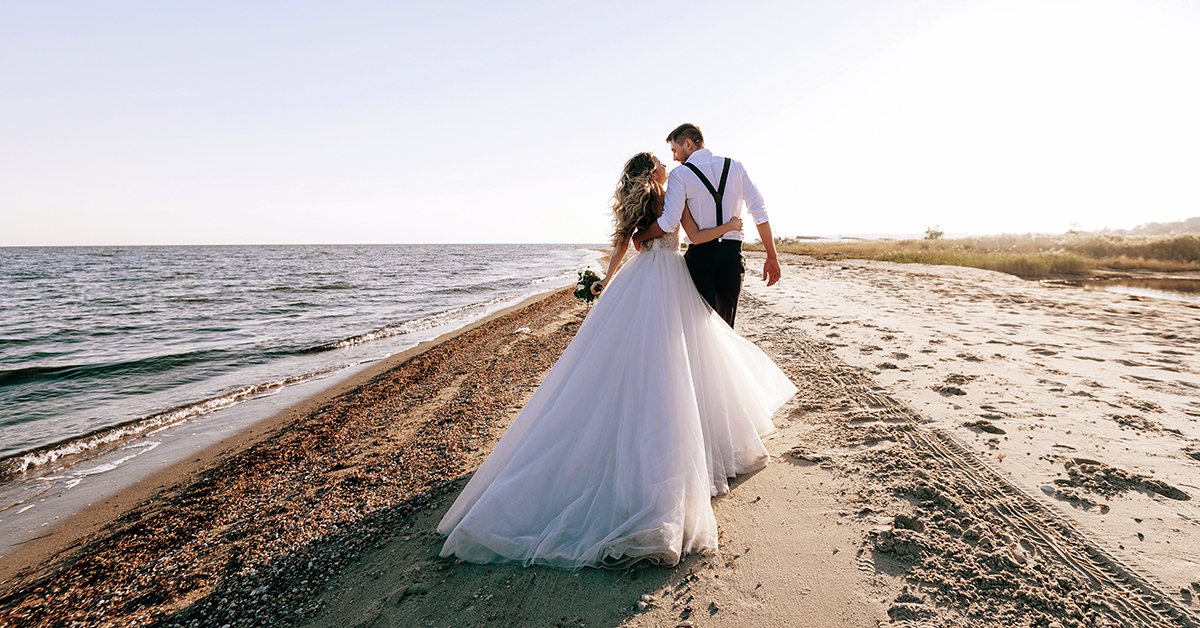 Couple in wedding attire walking along a sandy beach shoreline at sunset, with ocean waves on one side and a flowing white wedding gown trailing behind.