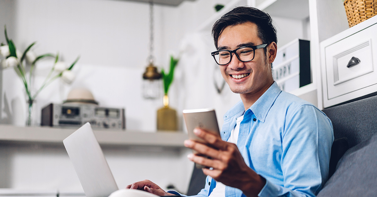 Teen smiling while checking his phone and using a laptop, representing the convenience of direct deposit for teens.