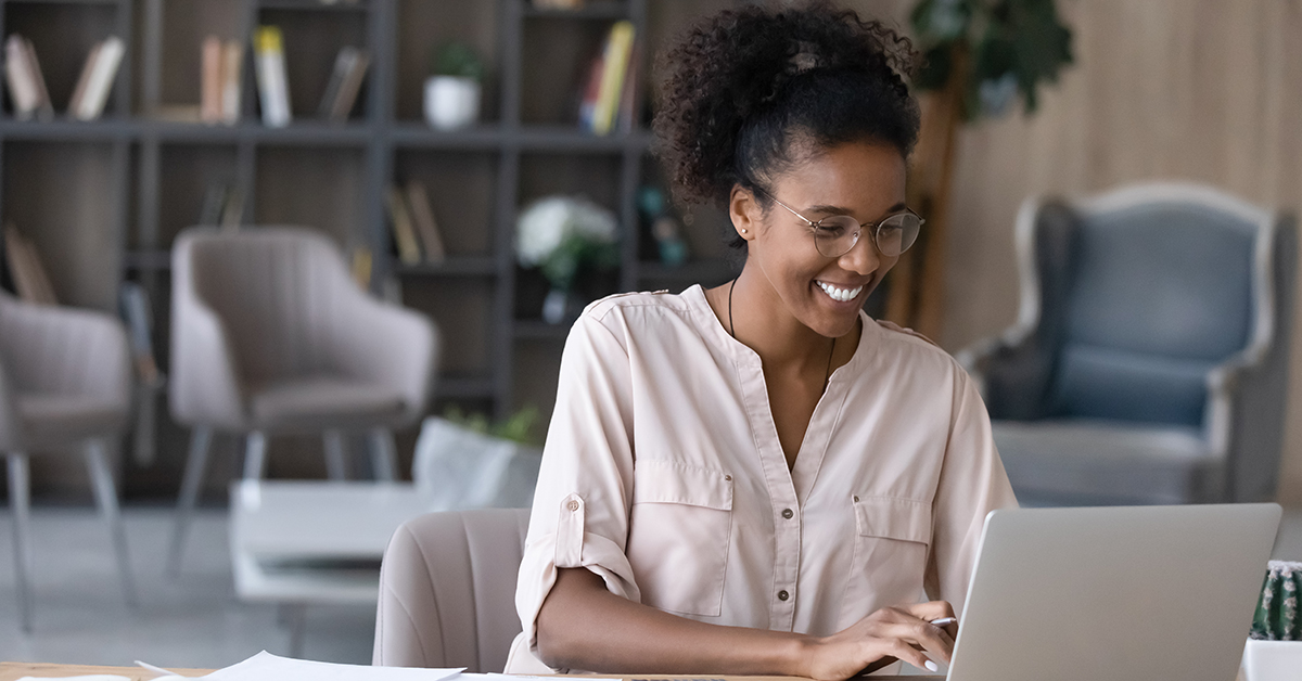 Smiling college student working on a laptop while reviewing financial documents in a modern lounge or library setting.
